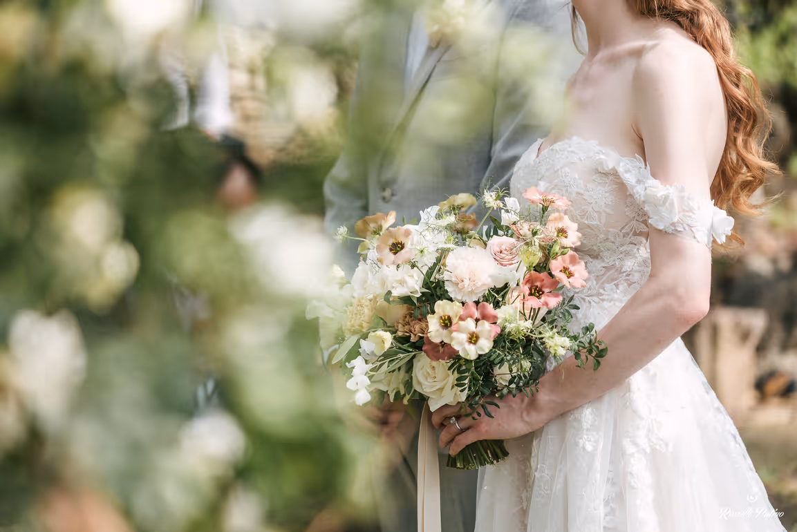A soft, artistic close-up of a bride in an off-the-shoulder lace wedding dress holding a pastel-toned floral bouquet, framed by blurry green leaves in a Tuscan garden.