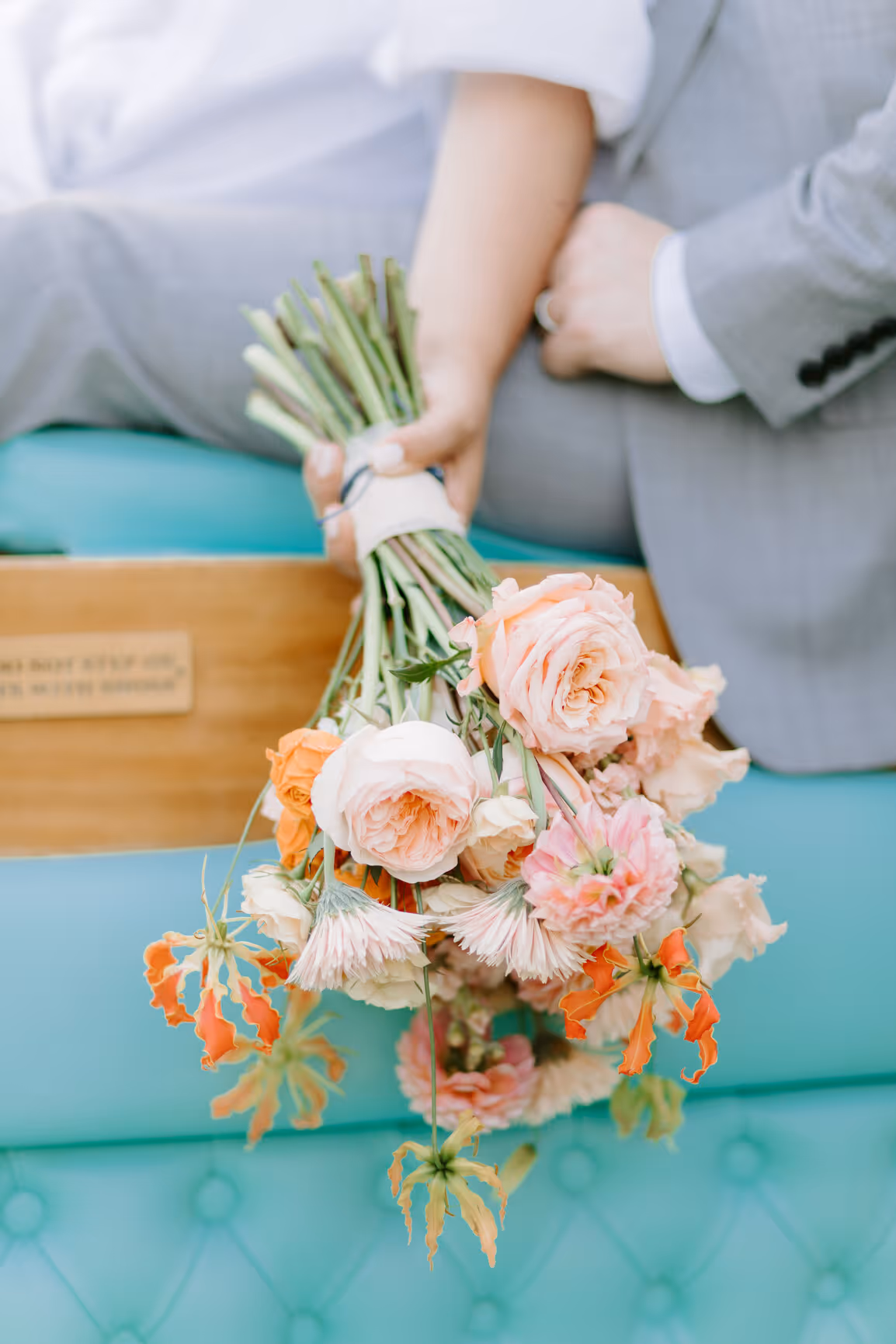 A vertical close-up detail shot of a bridal bouquet featuring peach garden roses and orange flowers held against the turquoise leather cushions of a classic Riva boat on Lake Como.