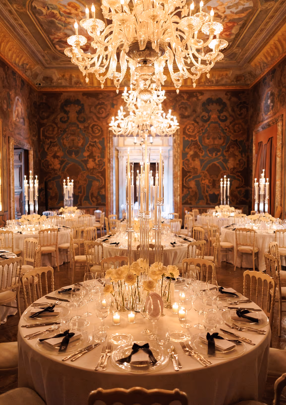 Spettacolare veduta del salone storico affrescato di Villa Erba allestito per un ricevimento di nozze di lusso, con imponenti lampadari di cristallo illuminati e tavoli tondi decorati con candelabri e candele.