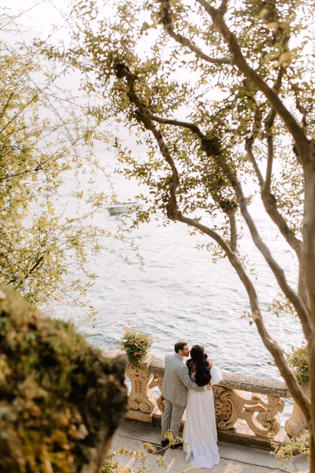 A vertical wedding photograph of a bride and groom kissing by the ornate stone balustrade at Villa del Balbianello, framed softly through the branches of lakeside trees overlooking Lake Como.