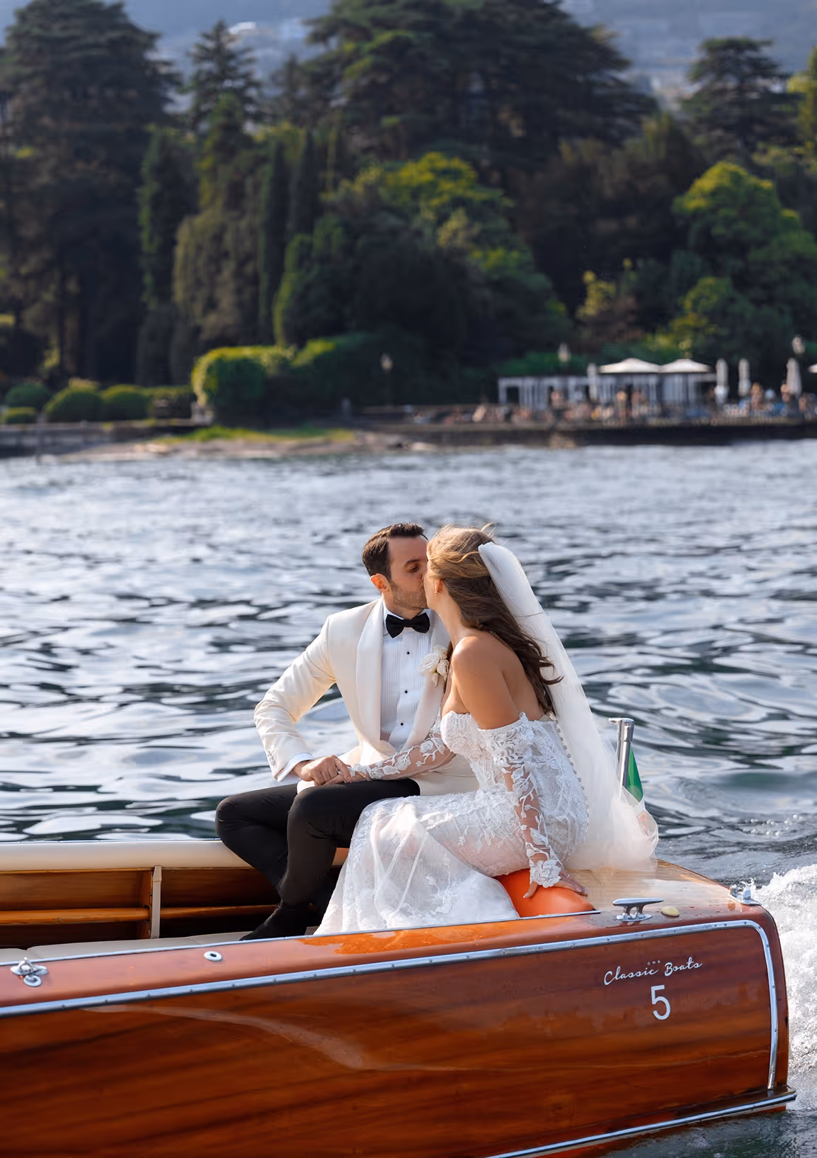 Primo piano a colori degli sposi mentre si baciano seduti a bordo di un motoscafo d'epoca in legno lucido, durante il loro destination wedding a Villa Erba sul Lago di Como.