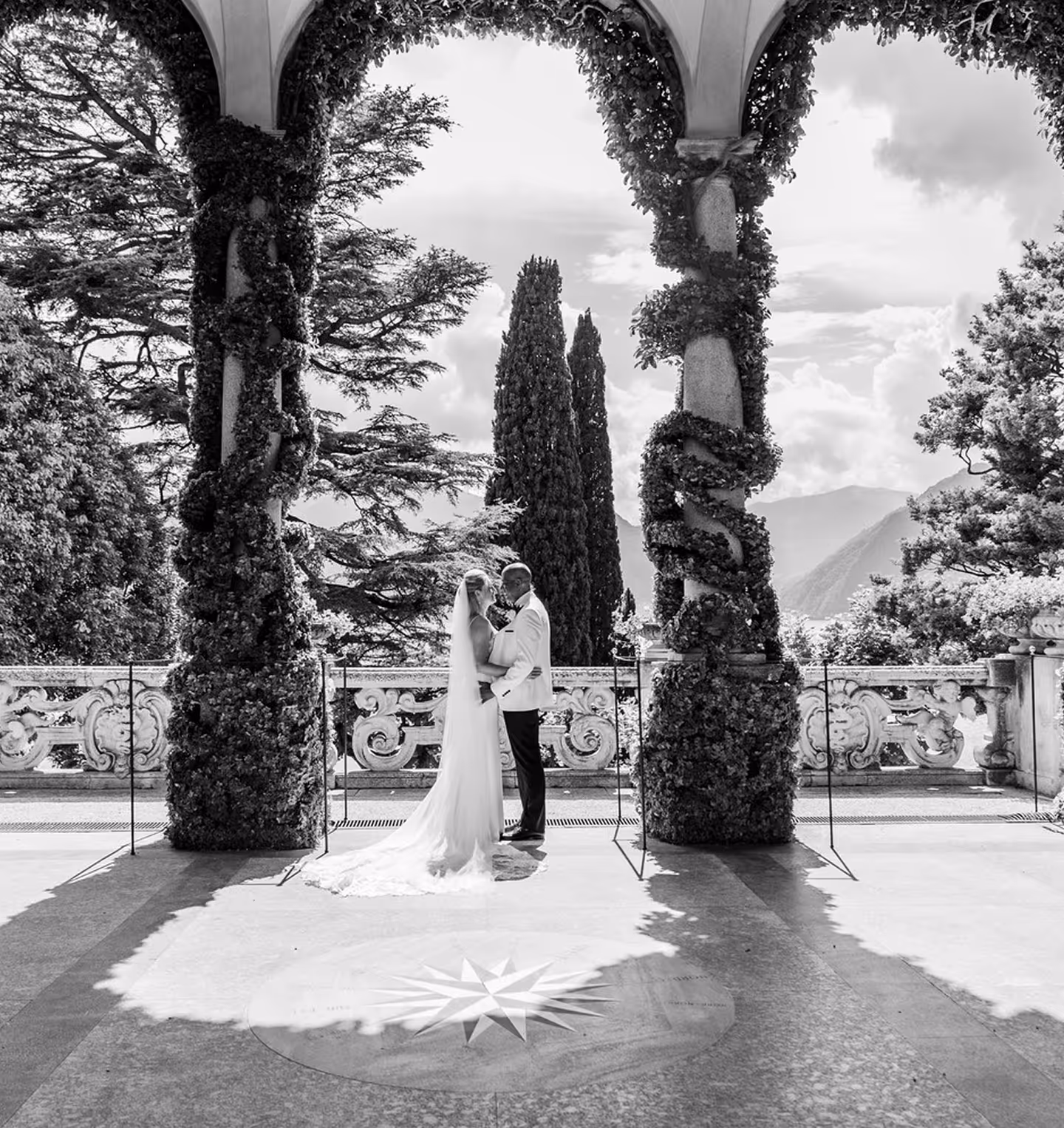 Foto in bianco e nero di una coppia di sposi che si abbraccia sotto le maestose arcate di un porticato storico ricoperto di piante rampicanti a Villa del Balbianello sul Lago di Como.