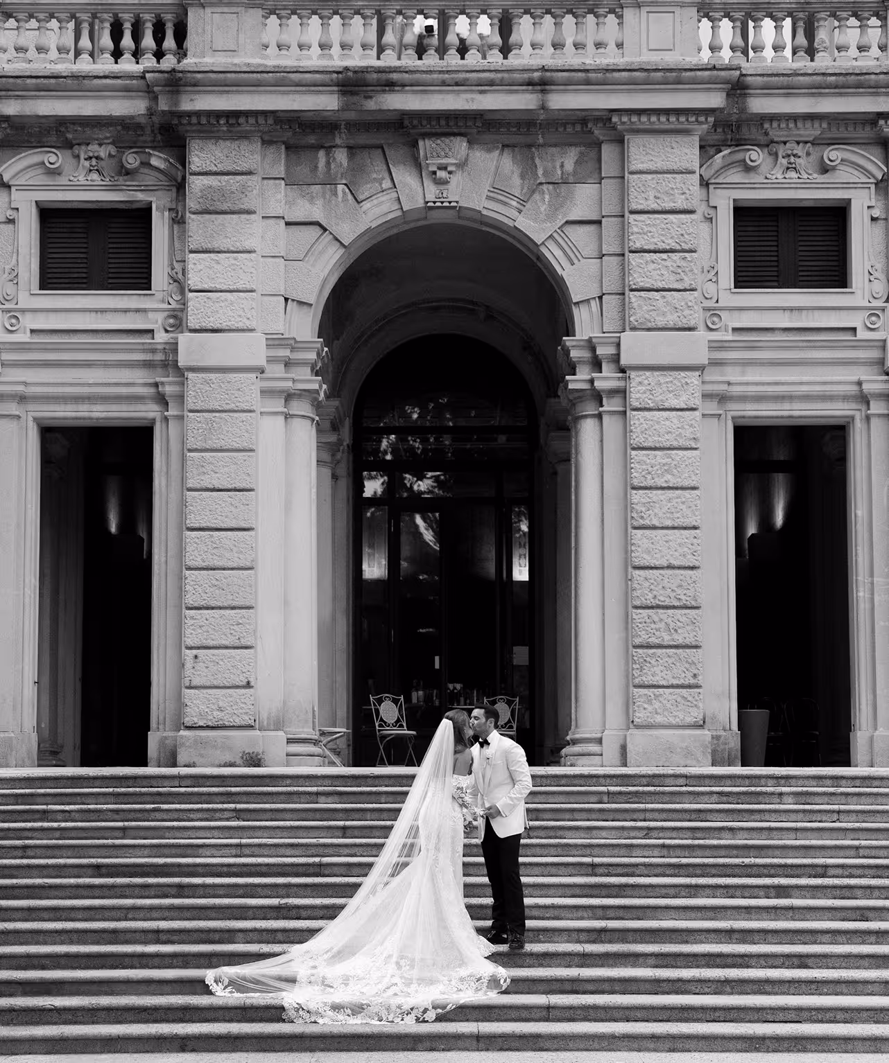 Foto in bianco e nero di una coppia di sposi che si bacia sulla monumentale scalinata in pietra all'ingresso di Villa Erba sul Lago di Como, con il lungo velo in pizzo della sposa adagiato sui gradini.