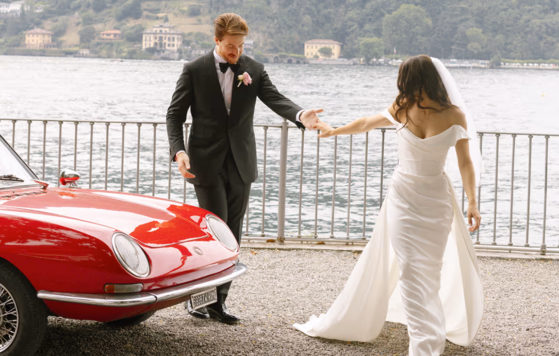 Lo sposo in smoking nero tiene per mano la sposa in abito bianco mentre camminano sul lungolago vicino a un'auto d'epoca rossa, per un matrimonio esclusivo a Villa del Grumello.