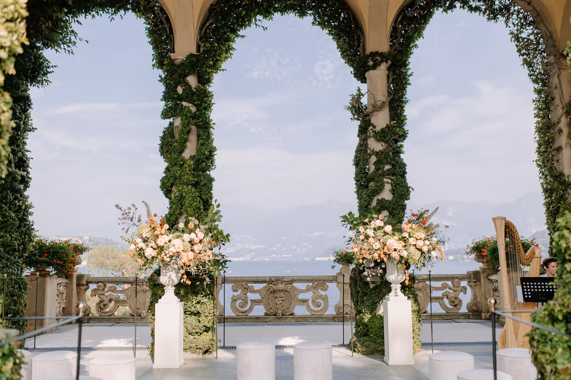An outdoor wedding ceremony setup at Villa del Balbianello on Lake Como, showing two ivy-covered stone pillars adorned with large peach floral arrangements, a classical balustrade, and a classical harp.