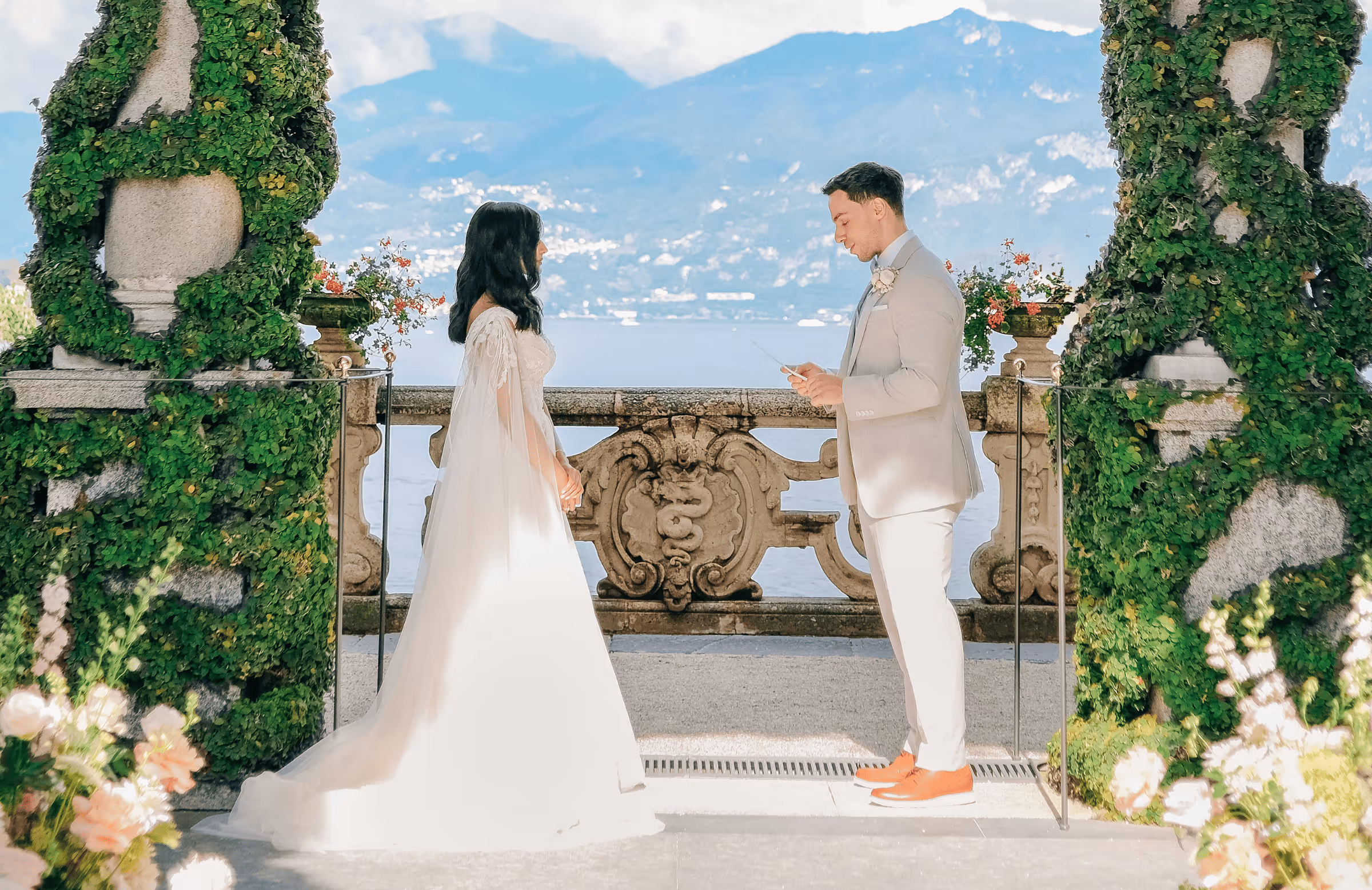 A landscape photograph of Puja and Rino exchanging vows during their wedding ceremony at Villa del Balbianello, framed by ivy-covered stone pillars against the scenic backdrop of Lake Como.