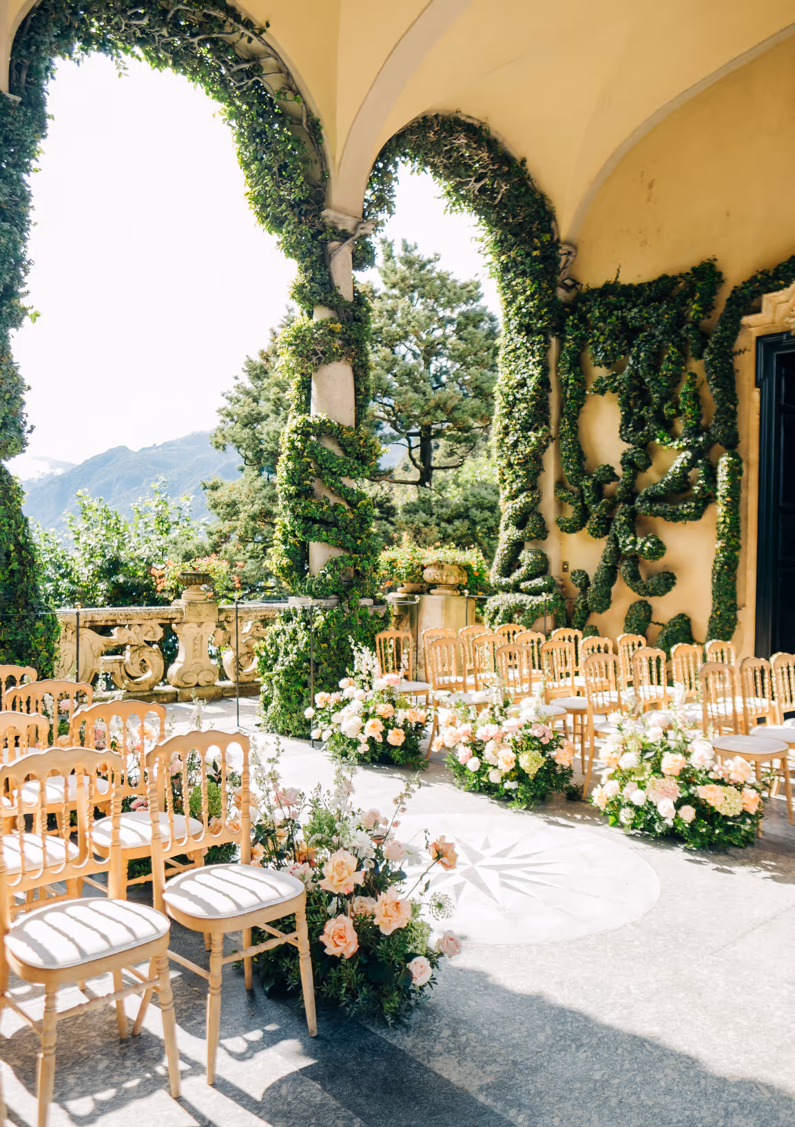 A sunlit vertical photograph of an outdoor wedding ceremony setup under the iconic ivy-draped arches of the loggia at Villa del Balbianello, featuring golden chairs and ground-level pastel floral arrangements.