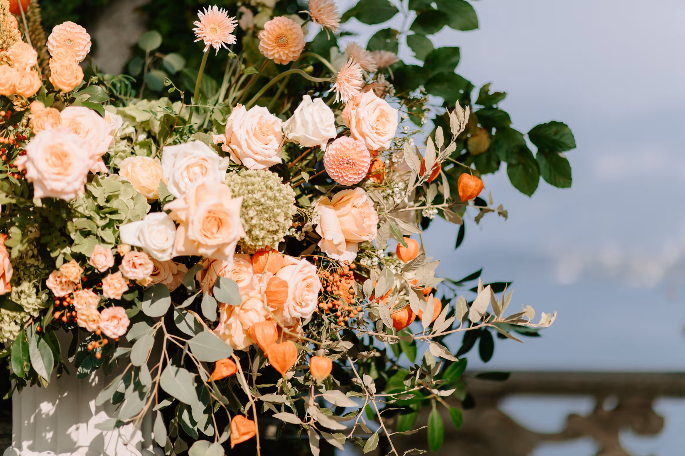 A close-up detail shot of a luxury wedding floral arrangement at Villa del Balbianello featuring peach roses, pink dahlias, and vibrant orange Chinese lantern pods overlooking Lake Como.