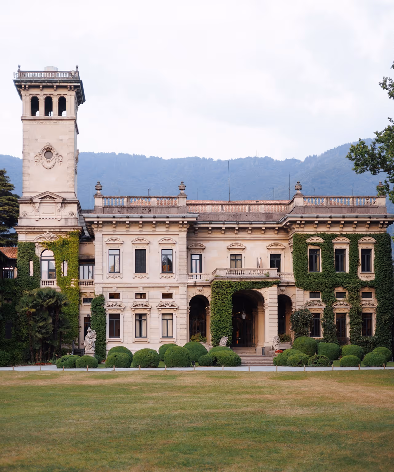 Una fotografia frontale della monumentale facciata di Villa Erba a Cernobbio, con la sua antica torre, l'edera rampicante e le siepi decorative geometriche nel prato antistante.