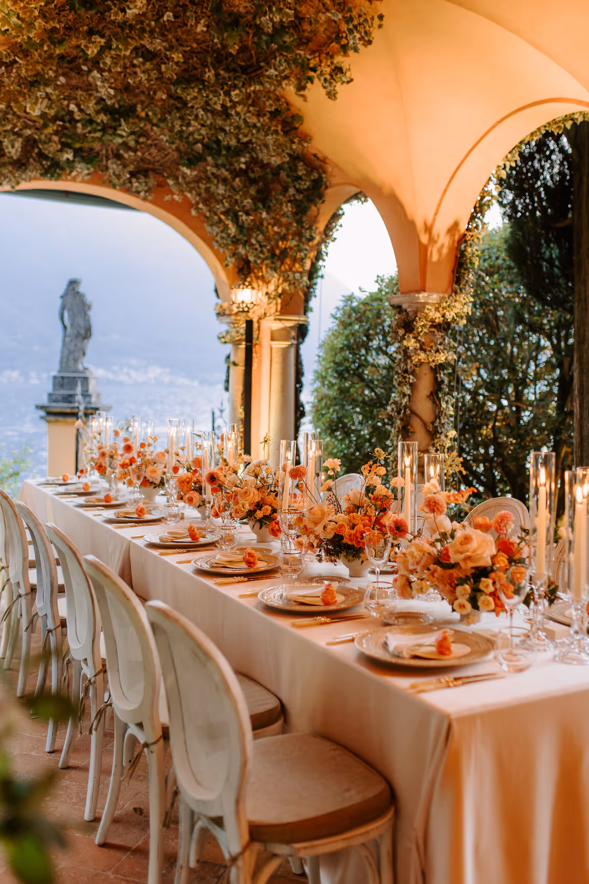 A luxury wedding reception dinner setup under the historic arched loggia at Villa La Cassinella, featuring a long table adorned with peach roses, terracotta florals, and glowing cylinder candles overlooking Lake Como.