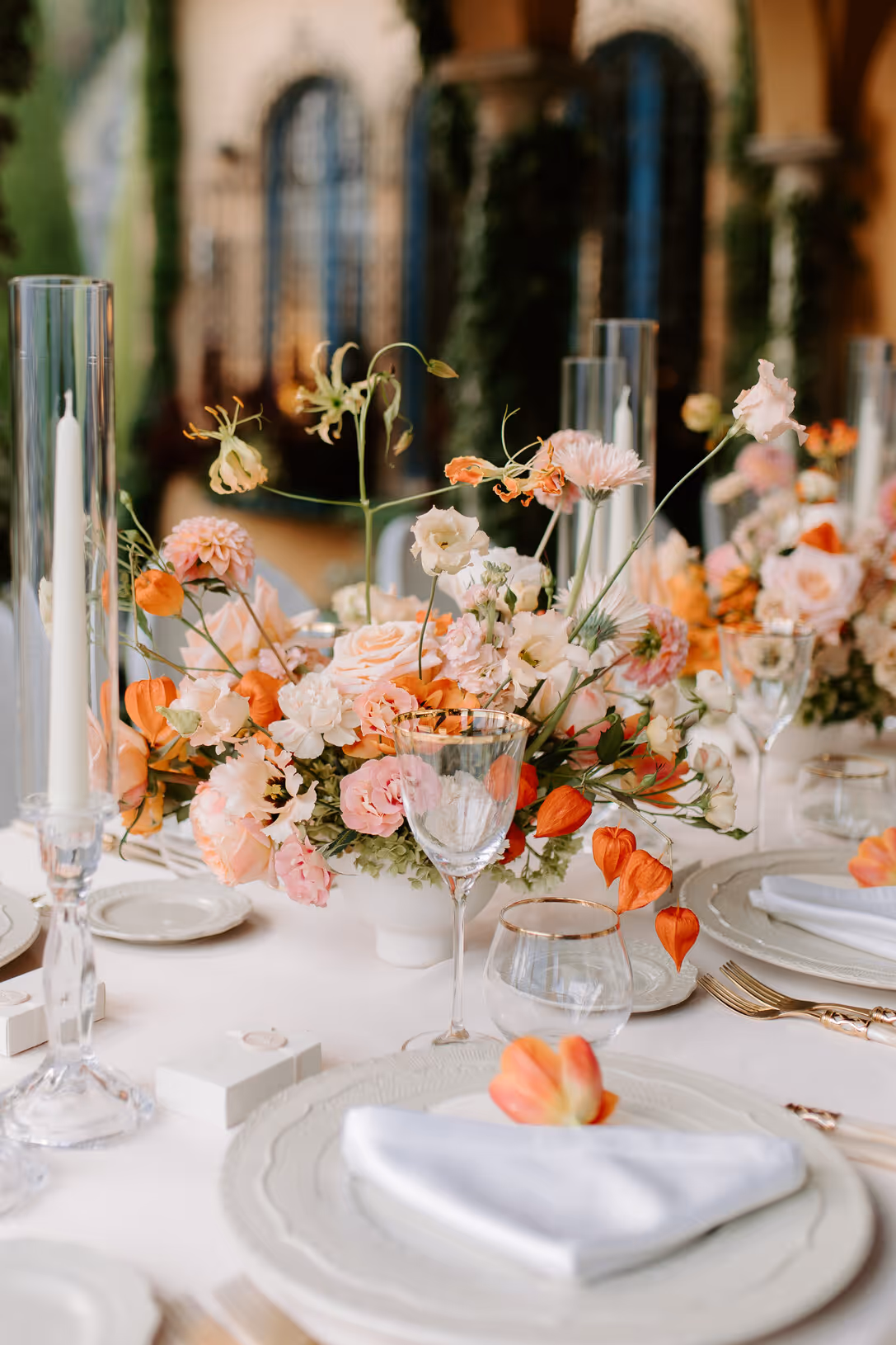 A close-up vertical photograph of a luxury wedding reception dinner table at Villa La Cassinella, detailed with white plates, gold flatware, tall glass candles, and a coral and peach floral centerpiece.