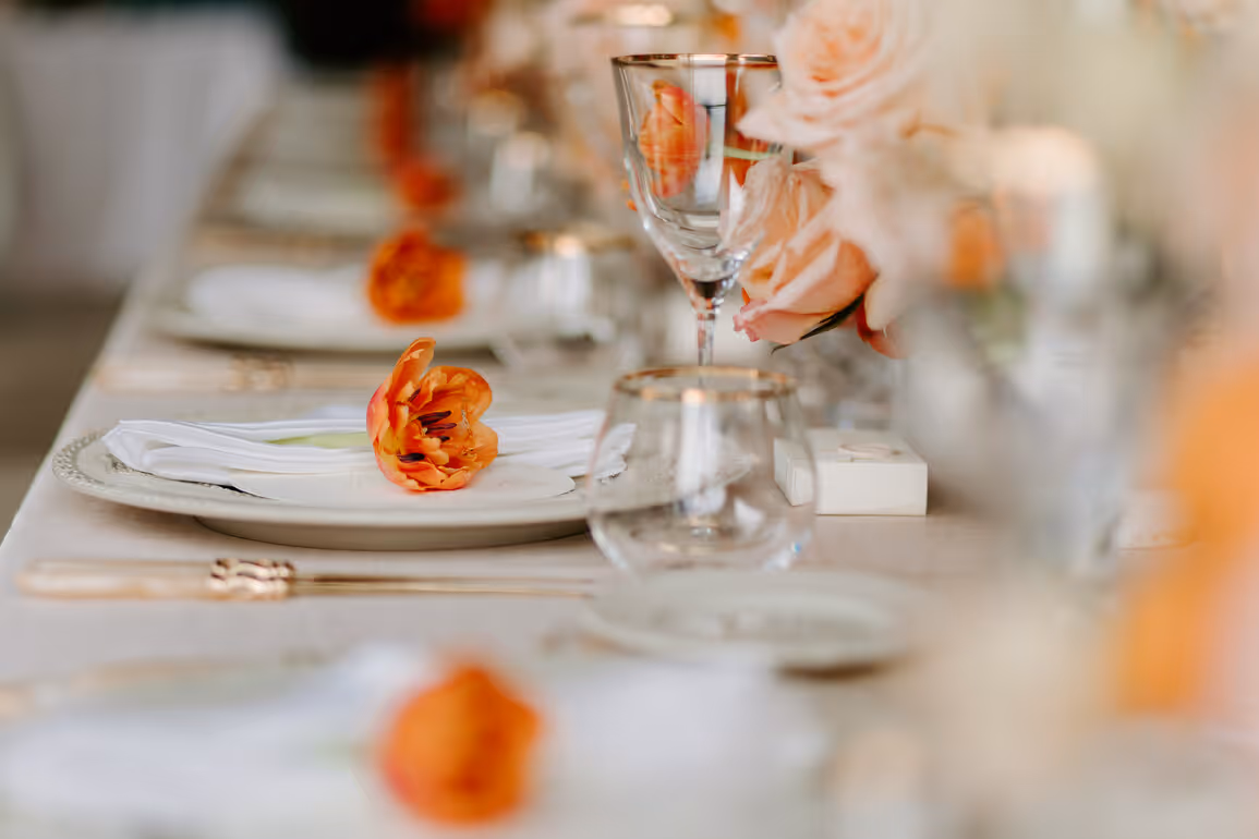 A close-up macro photograph of an elegant wedding table setting at Villa La Cassinella, featuring gold-rimmed crystal glassware, an orange tulip on a white linen napkin, and soft blush pink roses.