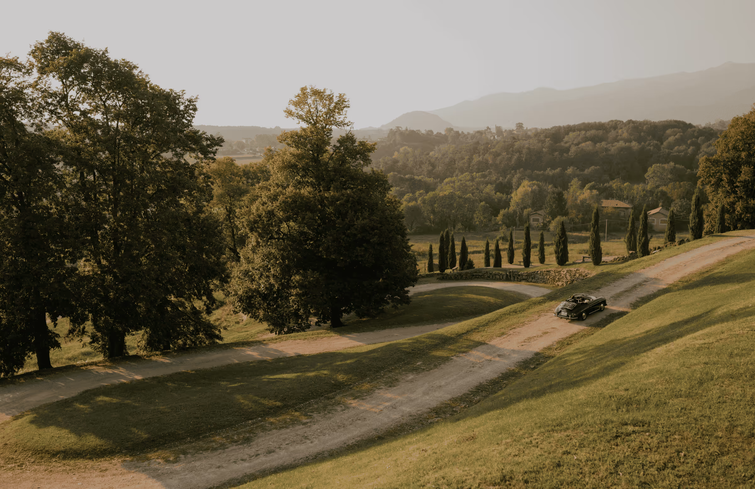 A cinematic wide landscape photograph of a black vintage convertible driving along a winding dirt road through the rolling green hills and cypress trees of the Italian countryside near Castello Durini.