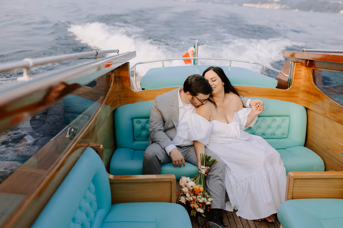A wide candid shot of a bride and groom relaxing on turquoise leather seats inside a luxury wooden water taxi on Lake Como, with the water wake visible in the background.
