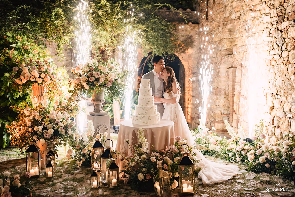 A festive nighttime photograph of a bride and groom smiling during their wedding cake cutting, illuminated by bright sparkler fountains against the rustic stone walls of a Tuscan castle.