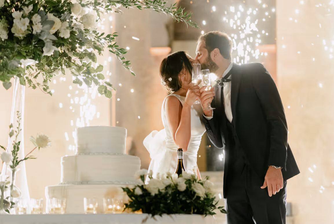 A dynamic reception photograph of Martina and Alessandro kissing and holding champagne flutes during their wedding cake cutting, framed by brilliant golden fountain sparklers.