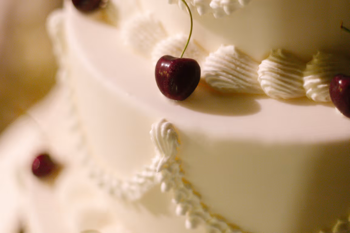 A macro detail shot of a classic white frosted wedding cake adorned with piped cream frosting decorations and a fresh dark cherry.